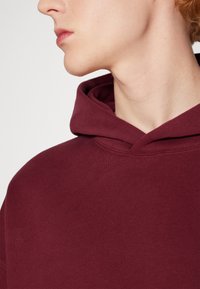 Sudadera color burdeos con bolsillo canguro delantero, capucha con cordón y puños acanalados. Fabricada en una suave mezcla de algodón.