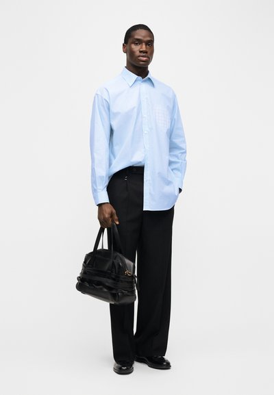 Άνδρας στέκεται φορώντας ανοιχτό μπλε πουκάμισο με κουμπιά, μαύρο παντελόνι και μαύρα παπούτσια, κρατώντας με τα δύο χέρια μια μαύρη δερμάτινη τσάντα.