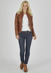 Braune Lederjacke mit Reißverschluss, gerippter Textur und seitlichen Taschen. Kombiniert mit einem weißen Oberteil, dunklen Jeans und braunen Stiefeletten.