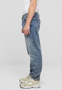 Blugi din denim albastru deschis, cu croi relaxat, textură estompată și cusături vizibile. Purtați cu adidași bej. Vedere din lateral.