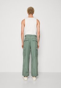 Cargo bukser i lys grøn stof med bred ben-design. Har to store sidelommer og en forstærket talje. Bæres med en hvid tank top.