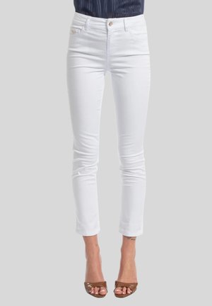 Pantaloni bianchi slim-fit cropped indossati con tacchi aperti marroni e top scuro infilato, mostrati dalla vita ai piedi.