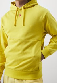 Sweat à capuche jaune en tissu doux, avec une poche kangourou à l'avant, des cordons de serrage et un petit logo sur la poitrine.