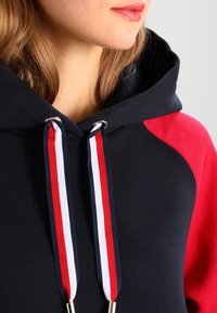 Marineblauer und roter Hoodie mit einer Kordel, der rot-weiße und marineblaue Streifen sowie goldfarbene Metallspitzen aufweist. Glatte Textur mit lässigem Design.