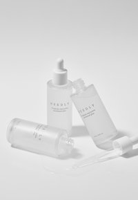 Three clear glass bottles with droppers, labeled "NEEDLY," containing a lightweight, clear liquid. One bottle is tipped, spilling product.