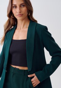 Zelený tailored blazer s širokým límcem, vyrobený z hladké tkaniny. Obsahuje přiléhavý střih, kombinovaný s černým cropped topem a odpovídajícími kalhotami.
