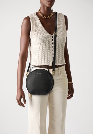 Schwarze Leder-Crossbody-Tasche mit runder Form, goldfarbenen Beschlägen und strukturierter Oberfläche. Getragen zu einem cremefarbenen ärmellosen Strickoberteil.
