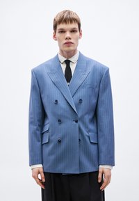 Sacou dublu cu dungi albastre, rever lat, două buzunare frontale și nasturi negri, purtat peste o cămașă albă și o cravată neagră.