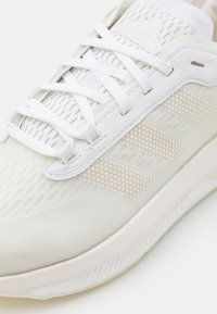 Zapatilla deportiva blanca con un upper texturizado, que presenta paneles de malla y rayas punteadas en contraste. Diseño de cordones con una suela acolchada.
