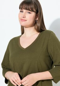 Olivově zelen�ý top s v-výstřihem a tříčtvrtečními rukávy, vyrobený z měkké látky. Vyznačuje se texturovaným povrchem a kontrastním detailem u výstřihu.