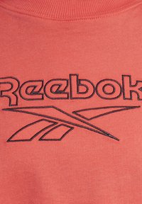 Korálově růžová mikina s logem "Reebok" v černé výšivce a elegantním liniovým designem. Měkká tkanina, žebrovaný výstřih a moderní střih.
