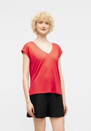 Γυναίκα με κοντά σγουρά ξανθά μαλλιά, φορούσα κόκκινο τοπ με V γιακά και κοντά μανίκια, και μαύρο σορτς, στέκεται μπροστά σε απλό φωτεινό φόντο.