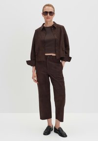 Braune Wildleder-Cropped-Jacke, braunes eng anliegendes Oberteil und passende gerade geschnittene Hose. Die Accessoires bestehen aus dunkler Sonnenbrille und schwarzen Lederloafers.