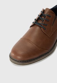 Chaussure en cuir marron avec un bout arrondi, un dessus texturé et des lacets sombres contrastants. Présente une semelle cousue et un minimum de détails.