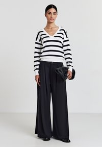 Woman standing, wearing a white sweater with black horizontal stripes, wide black pants, holding a black clutch bag against a plain background.