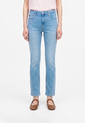 Kvinde iført lyseblå jeans med højt talje og lige ben, en sart lyserød ærmeløs top og brune flade sko, stående på en hvid baggrund.