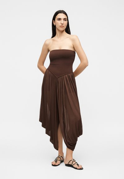 Γυναίκα με μακριά σκούρα μαλλιά που φοράει ένα στράπλες, καφέ, ασύμμετρο πλισέ φόρεμα και μαύρα σανδάλια με λουράκια, στέκεται με τα χέρια πίσω από την πλάτη.