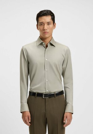 Chemise à manches longues gris clair avec des rayures subtiles, col classique et boutons blancs. Associée à un pantalon habillé texturé marron et une ceinture noire.