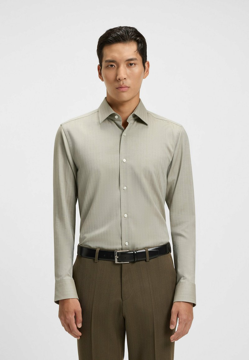 Camicia a maniche lunghe di un grigio chiaro con sottili righe, colletto classico e bottoni bianchi. Abbinata a pantaloni eleganti marroni con texture e una cintura nera.