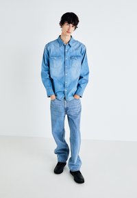 Camisa de denim en azul claro, con un diseño de botones y dos bolsillos en el pecho. Combinada con jeans holgados de azul claro y zuecos negros.