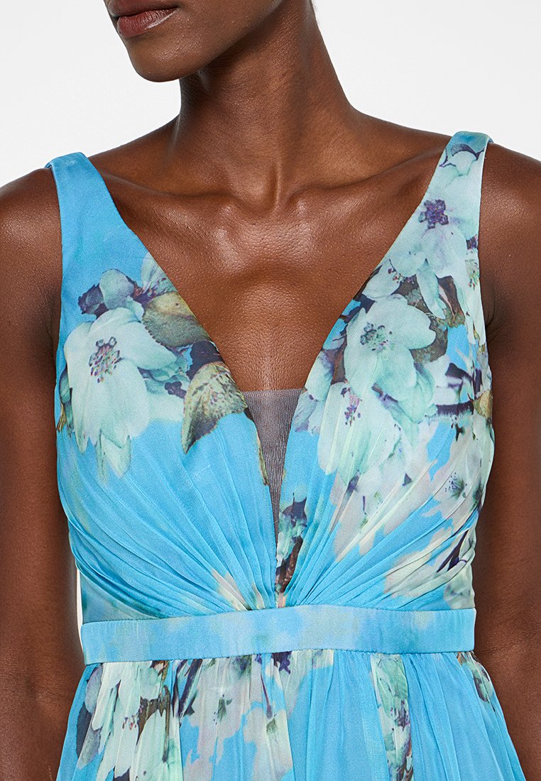 Helles blaues Blumenkleid mit tiefem V-Ausschnitt, gefaltetem Oberteil und einem strukturierten Stoff, der zarte weiße und grüne Blumenmuster zeigt.