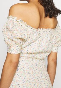 Kobieta w białej sukience odsłaniającej ramiona, ozdobionej małymi różowymi, fioletowymi i zielonymi wzorami kwiatowymi, pokazująca swoje plecy i prawą rękę.