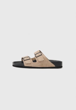 Suede čības ar gaiši bežīgu augšdaļu, melnu gumijas zoli un divām regulējamām sprādzēm drošai piestiprināšanai. Plakana un atvērta dizaina.