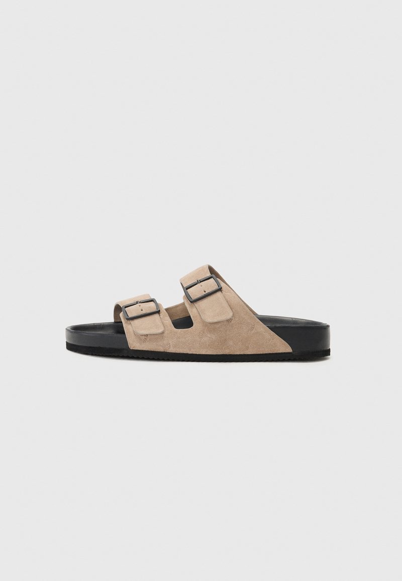 Suede sandaler med en lys beige overdel, sort gummisål og dobbelt justerbare spænder for en sikker pasform. Flad og åben design.