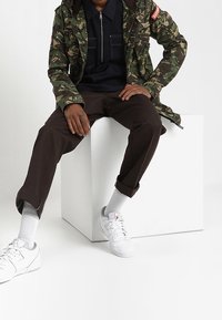 Chaqueta de camuflaje, polo oscuro con cremallera, pantalones marrones, zapatillas blancas y calcetines blancos visibles. Diseño simple y moderno con detalles prácticos.