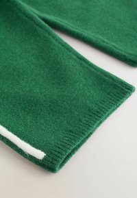 Zaļa adīta auduma ar mīkstu tekstūru, kam ir balta akcenta strīpa. Malas ir ribotas, nodrošinot pabeigtu izskatu.