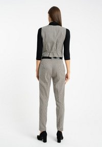 Kobieta w czarnej bluzce z rękawem 3/4 oraz szarej kamizelce w kratę, w dopasowanych spodniach, stoi do kamery tyłem na białym tle.