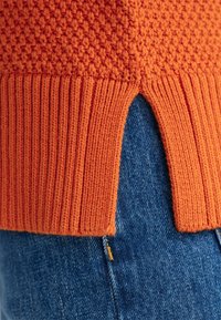 Oranger Strickpullover mit strukturiertem Oberteil und geripptem Saum, mit einem Schlitzdesign, das Denim darunter sichtbar macht.