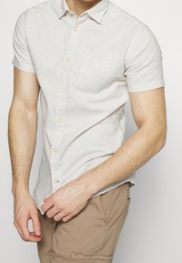 Homme portant une chemise à manches courtes beige clair avec de fines rayures horizontales et un pantalon beige, ajustant l'ourlet de sa chemise avec la main gauche.