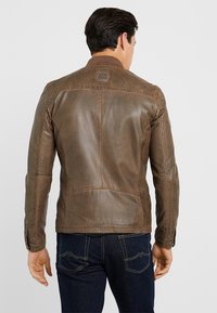Giacca in pelle marrone con finitura opaca, design aderente e colletto alto. Presenta dettagli di cucitura e un'etichetta con il marchio sulla schiena.