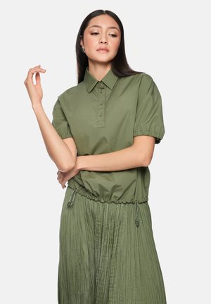 Woman with long dark hair wearing an olive green short-sleeve collared shirt and matching pleated skirt, posing with one arm raised.