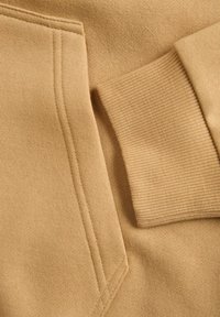 Tansweatshirtstof met een gladde textuur, voorzien van een ribgebreide manchette en een platte zakdetail, met schone stiksels en een eenvoudig ontwerp.