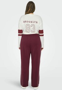 Burgundske sportske hlače širokog kroja s bijelim bočnim trakama, uparene s bijelom dugom majicom na kojoj se na leđima nalaze natpisi "BROOKLYN" i "83".