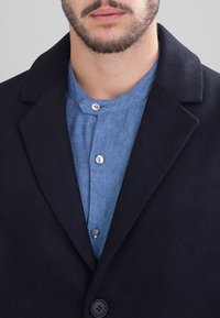 Cappotto in misto lana blu navy con rever a incavo e chiusura con un solo bottone, indossato sopra una camicia chambray azzurro chiaro con dettagli a bottoni.