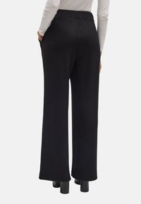 Pantalones negros de pierna ancha con una tela lisa, cinturilla elástica, bolsillos laterales y un dobladillo limpio. Mostrados desde la parte de atrás, combinados con tacones negros.
