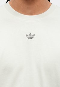 Svetlo šedé bavlnené tričko s malým, vyšitým čiernym logom Adidas na hrudi. Okružný výstrih s rebrovanou textúrou.