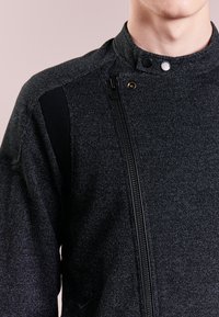 Giacca nera in tessuto con chiusura a zip e bottoni a pressione, caratterizzata da dettagli in tessuto nero a contrasto sulle spalle.