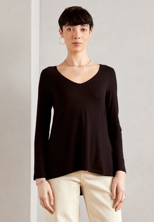 Black long-sleeve V-neck top made of soft fabric, features a relaxed fit with an asymmetrical hem, paired with light-colored, fitted pants.