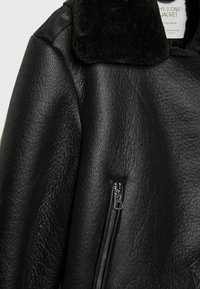 Giacca di pelle nera con superficie texturizzata, colletto foderato in pelliccia e tasca con zip sulla manica sinistra.