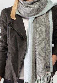 Écharpe bleu clair et grise à motif floral, texture légère et bords frangés, portée avec une veste sombre.