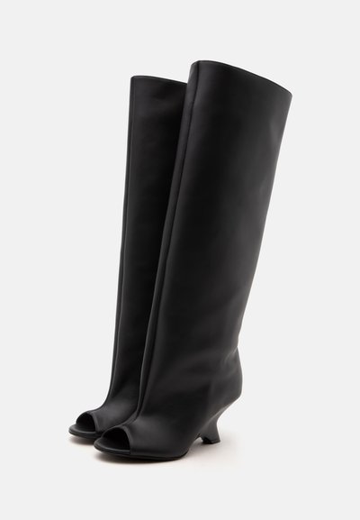 Bottes hautes en cuir noir avec bouts ouverts et petits talons bas présentées sur un fond uni.