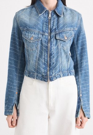 Person trägt eine blaue Jeansjacke mit Reißverschluss, Brusttaschen und langen Ärmeln, kombiniert mit weißen, hoch taillierten Hosen.