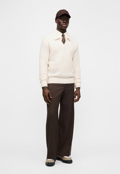 Cream textured sweater, brown wide-leg trousers, brown cap, and patterned tie. Footwear features a cream and black design. Simple and modern profile.