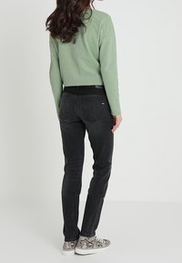 Maglione verde texturizzato, jeans slim-fit neri e sneaker a fantasia. Tasche posteriori visibili sui jeans, look complessivo casual.