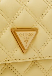 Gul quiltad faux läderhandväska med en triangulär guldsymbolplatta som har "GUESS U.S.A." och en frågeteckendesign.