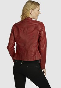 Rote Lederjacke mit einer taillierten Passform, die einen Stehkragen und Nähte aufweist. Kombiniert mit schwarzen Hosen, die einen Reißverschlussakzent zeigen.